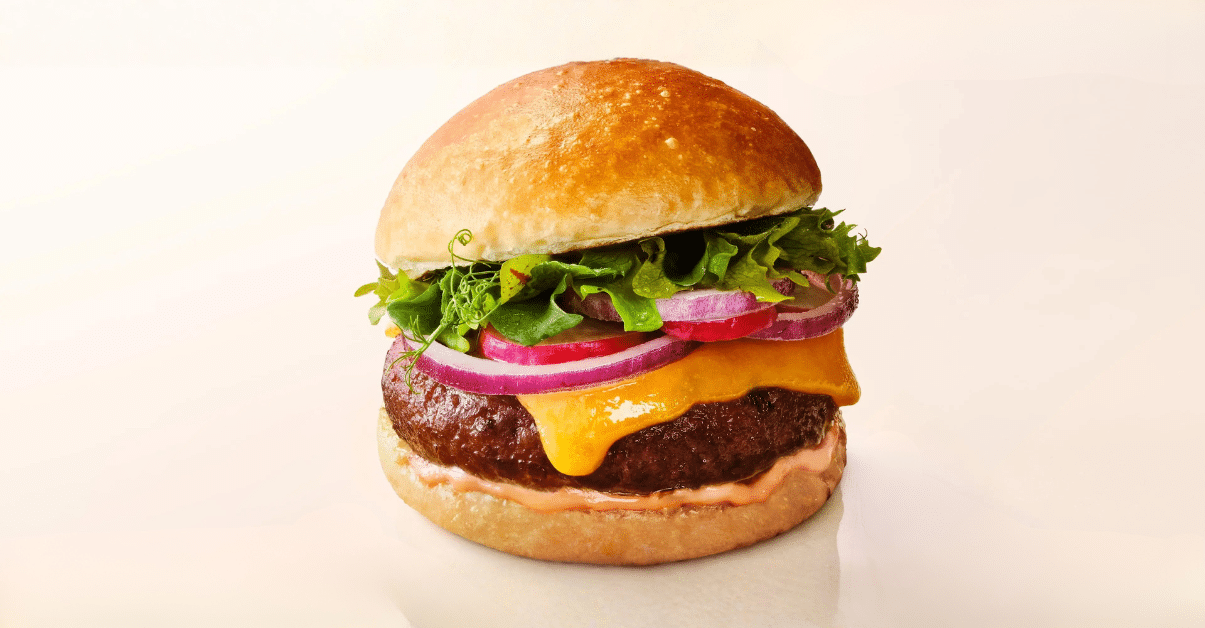 An image of a beef burger with cheese, onion, and lettuce on a plain beige background.