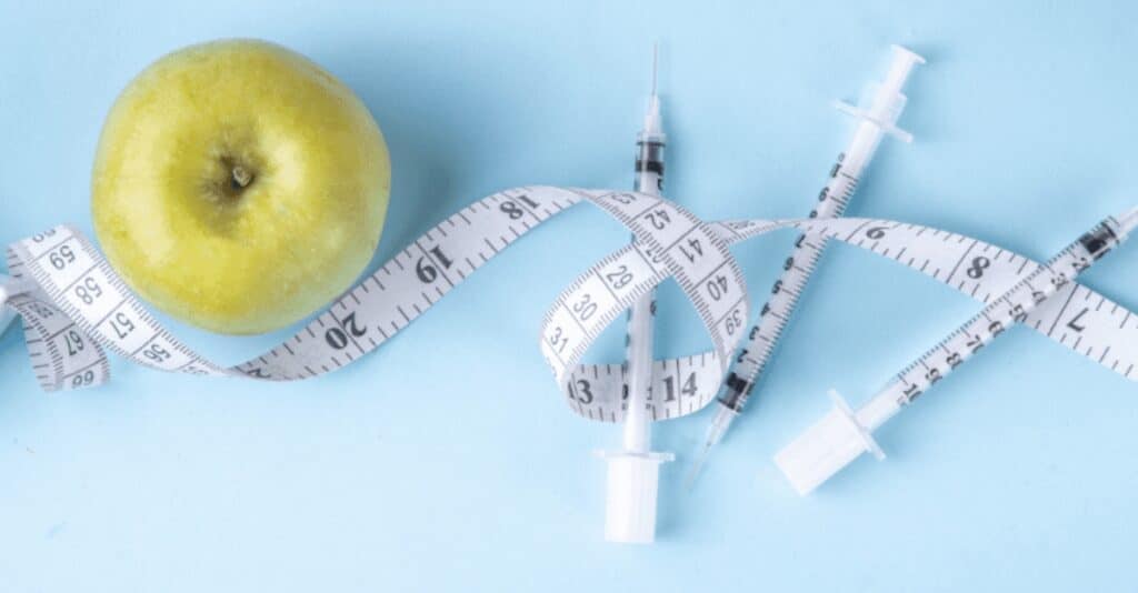 An apple sits in the top left of the image above a measuring tape that winds across the image, intertwining with three syringes.